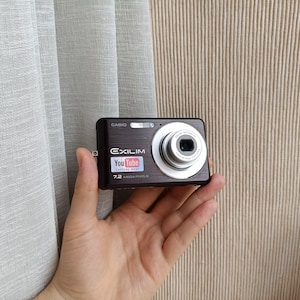 Puede incluir: Una cámara digital Casio Exilim de color marrón oscuro con un objetivo plateado, flash integrado y un logotipo de YouTube Capture Mode en la parte frontal. El cuerpo de la cámara muestra el texto Casio, Exilim y 7.2 Megapixels en blanco.