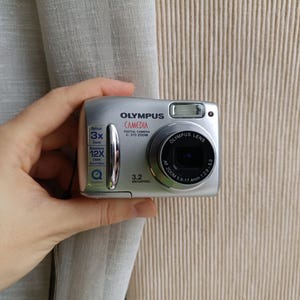 以下が含まれることがあります： シルバーのオリンパスCamediaデジタルカメラ。ズームレンズとフラッシュ付き。3.2メガピクセルの解像度で、3倍光学ズームと12倍デジタルズームを搭載。カメラは手で持たれています。