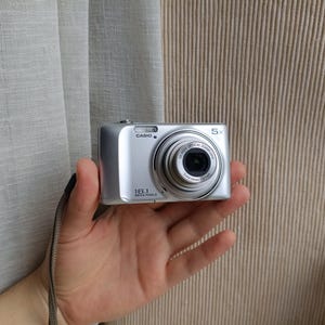 Pode incluir: Uma câmara digital Casio prateada com uma lente de zoom ótico de 5x, um sensor de 16,1 megapíxeis e uma correia de pulso cinzenta. A parte frontal da câmara apresenta a marca Casio e especificações técnicas impressas na lente.