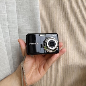 May include: A black Fujifilm digital camera with a silver lens barrel, featuring a 10.2 megapixels label on the front, a small built-in flash, and a light-coloured woven wrist strap attached to the side of the compact rectangular body.