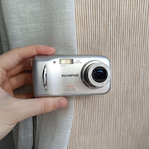 Op de afbeelding: Zilveren Olympus C-480 Zoom digitale camera met een resolutie van 4,0 megapixels. De camera heeft een lens met een zoombereik van 6,3-18,9 mm en wordt door een hand vastgehouden. De camera staat tegen een beige achtergrond.