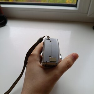 May include: A silver digital camera viewed from the side, featuring a metallic body, a small beige port cover, visible screws, and a brown fabric wrist strap attached to the top corner of the device casing.