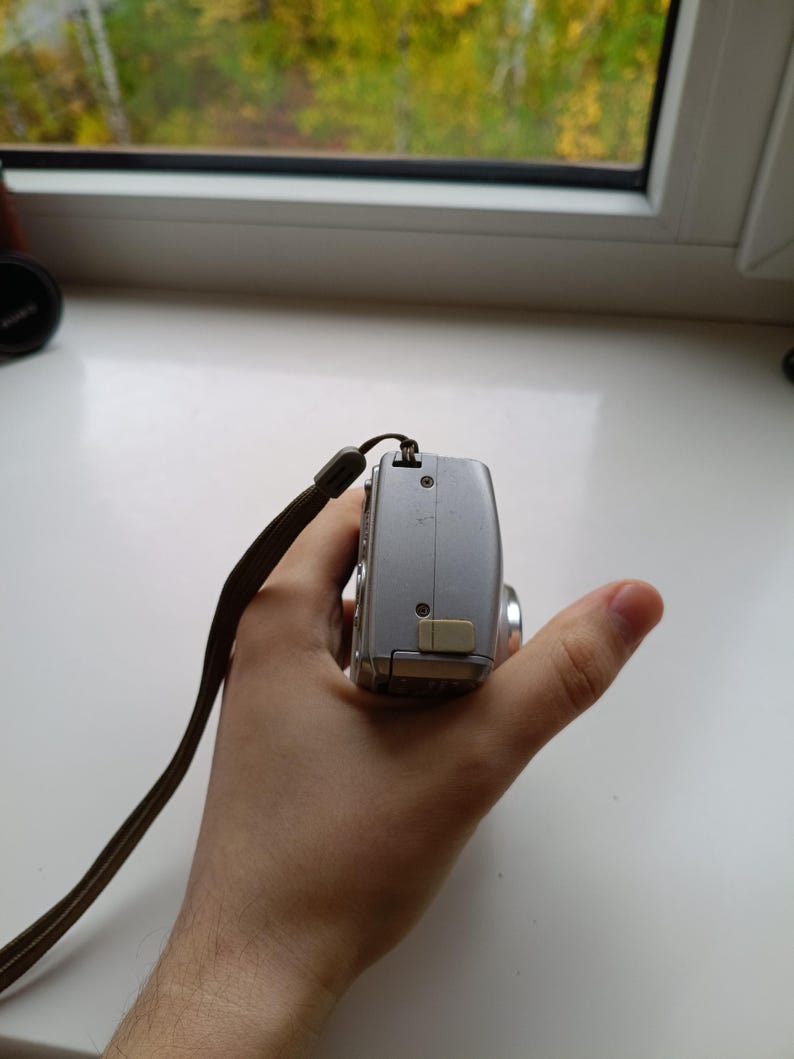 May include: A silver digital camera viewed from the side, featuring a metallic body, a small beige port cover, visible screws, and a brown fabric wrist strap attached to the top corner of the device casing.