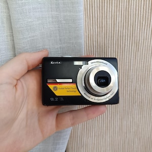 Könnte beinhalten: Eine schwarze Kodak Digitalkamera mit silbernem Objektiv und Blitz. Die Kamera hat einen gelb-schwarzen Aufkleber mit der Aufschrift "Kodak Perfect Touch technology" und "9.2". Die Kamera wird in einer Hand gehalten.