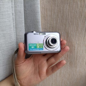 Puede incluir: Una cámara digital Fujifilm plateada con correa de muñeca. La parte frontal presenta un objetivo, un flash y una pegatina con el texto 16 Mega Pixels, Digital Image Stabilization, Smile & Blink Detection y Tracking Auto Focus.