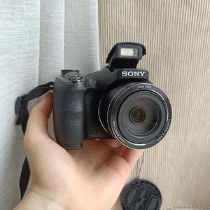 Puede incluir: Una cámara digital Sony negra con flash emergente, empuñadura texturizada y un objetivo con zoom óptico de 35x. La cámara incluye una correa negra para el cuello y una tapa de objetivo separada, con la marca Sony visible en la parte frontal.