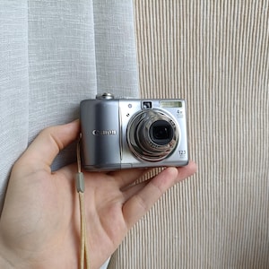 以下が含まれることがあります： 4倍光学ズームレンズ、内蔵フラッシュ、12.1メガピクセルセンサーを搭載したシルバーのキヤノン製デジタルカメラ。コンパクトな本体にはリストストラップが付いており、金属製のボディの左側にはキヤノンのロゴが表示されています。