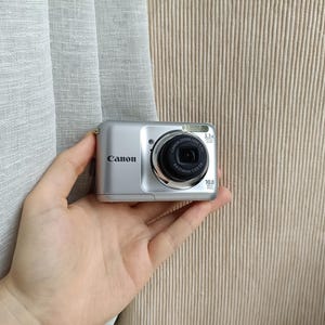 以下が含まれることがあります： 黒いレンズ鏡筒、内蔵フラッシュ、3.3倍光学ズームレンズを備えたシルバーのキヤノン製デジタルカメラ。カメラ本体の前面にはキヤノンロゴが表示され、レンズハウジングには10.0メガピクセルおよびレンズの仕様が記載されています。