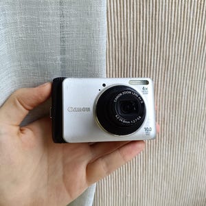 May include: Silver Canon digital camera with a black lens and a 4x optical zoom. The camera has a 10.0 megapixel resolution and a 6.2-24.8mm lens. The camera is held in a hand.