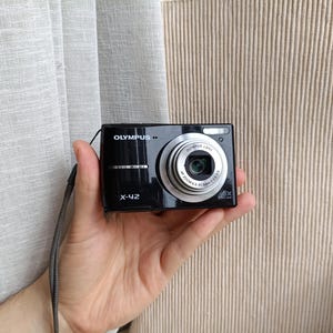 Puede incluir: Una cámara digital Olympus X-42 de color negro con un barril de lente plateado. La parte frontal de la cámara muestra la marca Olympus, el número de modelo X-42 y una lente con especificaciones de zoom óptico 6x alrededor del cristal.
