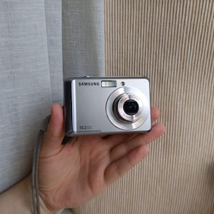May include: A silver Samsung digital camera featuring a 10.2 megapixel sensor, a built-in flash, and a lens with 3x optical zoom. The compact device includes a wrist strap attached to the side and displays the brand name on the front.