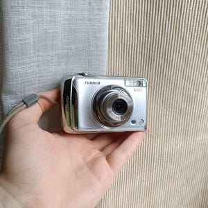 May include: A silver Fujifilm digital camera with a retractable lens and a built-in flash. The camera has a wrist strap attached and is held in a hand. The camera has the text "Fujifilm" printed on the top.