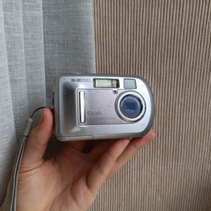 Puede incluir: Una cámara digital Kodak EasyShare de color plateado con un objetivo de 3,2 megapíxeles, flash integrado y un diseño de cuerpo rectangular. La parte frontal muestra la marca Kodak e incluye una correa de muñeca en el lateral.