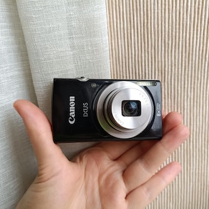 以下が含まれることがあります： 黒いCanon IXUSデジタルカメラ。シルバーレンズと8倍ズーム。カメラは手で持たれており、背景はニュートラルです。