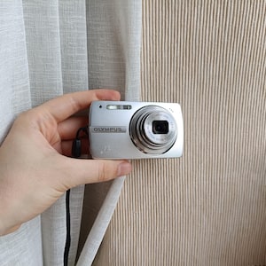 May include: A silver Olympus digital camera featuring a compact rectangular body, a prominent circular lens barrel with 5x optical zoom text, a built-in flash, and a black wrist strap attached to the side of the device casing.