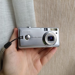 以下が含まれることがあります： ズームレンズ付きのシルバーのCanon PowerShot A400デジタルカメラ。カメラには黒いリストストラップとシルバーのボタンが付いています。カメラは手で持たれています。