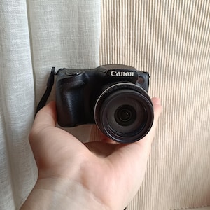 以下が含まれることがあります： 黒いCanonデジタルカメラ、レンズ付き、手に持たれています。カメラには黒いストラップがあり、Canonのロゴが見えます。背景にはベージュのカーテンとベージュの生地が含まれています。