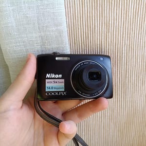 May include: A black Nikon Coolpix digital camera with a wide 5x zoom lens. The camera has a 14.0-megapixel resolution and a built-in flash. The camera is held in a hand, with a black strap attached.