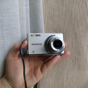 May include: A white Olympus FE digital camera with a silver lens barrel, a built-in flash, and a black wrist strap. The front of the camera features the Olympus brand name in silver lettering and the model designation fe in a cursive font.