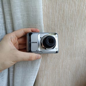 以下が含まれることがあります： 黒いレンズ、内蔵フラッシュ、10.0メガピクセルのセンサーを備えたシルバーのキヤノン製デジタルカメラ。カメラの前面にはキヤノンのロゴ、3.3倍光学ズームのラベル、焦点距離6.2-20.4mm、f/2.8-5.8のレンズが搭載されています。