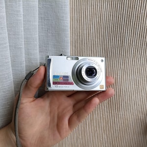 Puede incluir: Una cámara digital plateada Lumix con lente Leica, correa de muñeca y una etiqueta frontal que indica 10 Mega Pixels. El cuerpo de la cámara muestra varios iconos y textos técnicos, incluyendo Mega O.I.S. y Wide Angle.