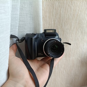 Puede incluir: Una cámara digital negra con un objetivo de zoom óptico de 10x, un flash emergente en la parte superior y una etiqueta de 6.0 megapíxeles. El dispositivo incluye una correa de cuello de tela negra unida al lateral y una empuñadura texturizada.