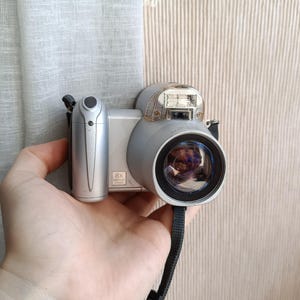 May include: A silver digital camera with a black strap is held in a hand. The camera has a large lens and a built-in flash. The lens is labeled with "8X". The camera is held in front of a beige wall and a white curtain.