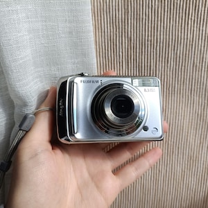 May include: Silver Fujifilm digital camera with a retractable lens and a built-in flash. The camera has a wrist strap attached and features the text "FUJIFILM" and "8.3 MEGA PIXELS".