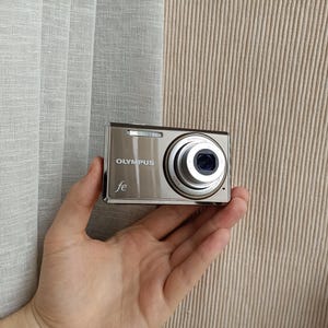 May include: A compact Olympus FE digital camera with a metallic silver finish. The front of the device features the Olympus brand name in white text, a small fe logo, a rectangular flash unit, and a circular lens barrel with silver detailing.
