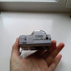 May include: A silver Nikon Coolpix digital camera viewed from the bottom, showing the battery and memory card compartment door, a tripod mount, and a label with the serial number 60327259 and Nikon Corp Japan text printed on it.
