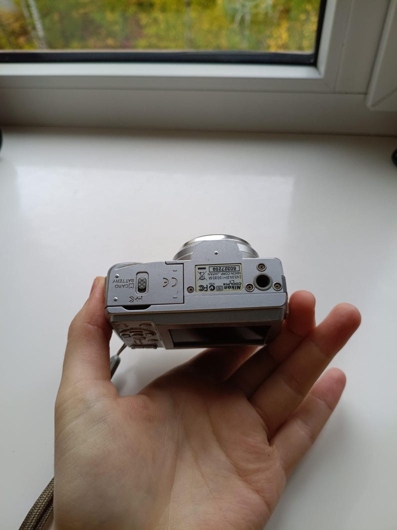 May include: A silver Nikon Coolpix digital camera viewed from the bottom, showing the battery and memory card compartment door, a tripod mount, and a label with the serial number 60327259 and Nikon Corp Japan text printed on it.