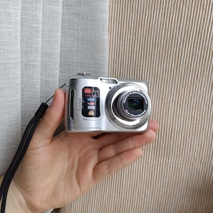 Könnte beinhalten: Eine silberne Kodak Digitalkamera mit einem 14-Megapixel-Sensor und einem 5-fach optischen asphärischen Objektiv. Auf der Vorderseite befinden sich der Markenname Kodak, ein kleiner Aufkleber mit Social-Media-Symbolen und eine schwarze Handschlaufe.