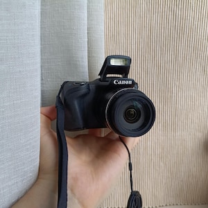 Puede incluir: Una cámara digital Canon negra con flash emergente, un objetivo circular prominente y una empuñadura texturizada. La cámara incluye una correa de muñeca negra unida al lateral y una tapa de objetivo sujeta por un cordón fino que cuelga.