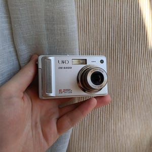 Op de afbeelding: Een zilveren UFO DS 6333 digitale camera met een 5,2 megapixel lens. De camera heeft een zoomlens en een klein LCD-scherm. De camera wordt in een hand gehouden.