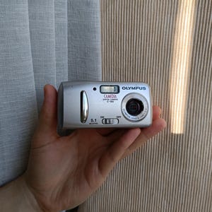 May include: A silver Olympus Camedia C-160 digital camera with a 5.1 megapixel sensor. The front of the device features a lens, a flash unit, an on-off power switch, and a vertical silver accent bar. The camera body is compact and rectangular.