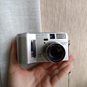 Può includere: Una fotocamera digitale argentata Minolta DiMAGE S414 con un obiettivo nero è tenuta in mano. La fotocamera ha un flash integrato e l'obiettivo legge 7,5-28,0 mm 1:3,0-3,9 ASPH. La fotocamera è da 4,0 megapixel.