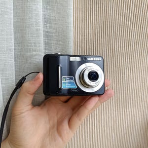 May include: A black Samsung D860 digital camera featuring a silver lens barrel, a built-in flash, and a rectangular body. The front of the camera displays a blue and white sticker indicating face detection and image stabilization features.