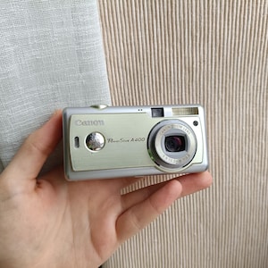 以下が含まれることがあります： ズームレンズ付きのシルバーのCanon PowerShot A400デジタルカメラが手に持たれています。カメラは、黒いレンズとフラッシュが付いたシルバーのボディをしています。背景は、ベージュの縦縞の生地です。