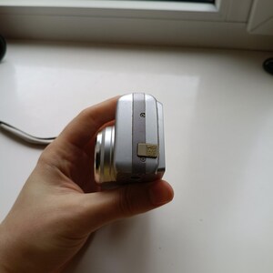 May include: A silver digital camera viewed from the side, featuring a metallic body, a protruding lens barrel, and a small beige rubber port cover labeled AV OUT with a USB icon, secured by two visible screws on the side panel.