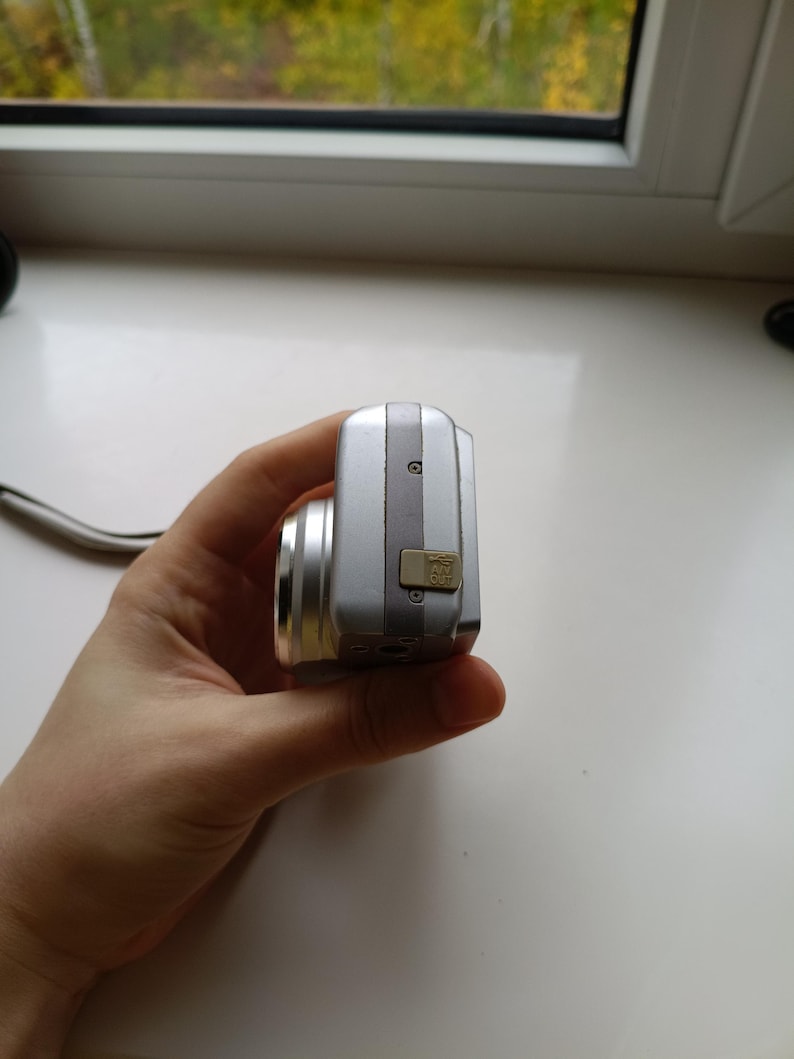 May include: A silver digital camera viewed from the side, featuring a metallic body, a protruding lens barrel, and a small beige rubber port cover labeled AV OUT with a USB icon, secured by two visible screws on the side panel.