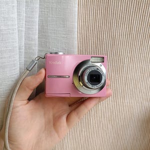Pode incluir: Uma câmara digital Kodak cor-de-rosa com uma lente prateada, flash integrado e um botão de disparo prateado na parte superior. Uma correia de pulso de cor clara está presa à lateral do corpo da câmara, que exibe a marca Kodak.