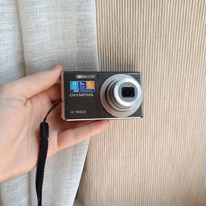 May include: A dark gray Olympus X-960 digital camera with a silver lens and a black wrist strap. The camera's screen displays blue and orange information. The camera is held in a hand.