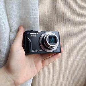 May include: A black Casio Exilim digital camera with a silver lens. The camera has the text "14.1" and "10x" printed on it. The camera is held in a hand.