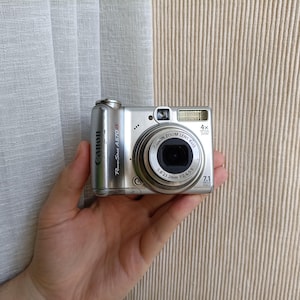 Puede incluir: Una cámara digital Canon PowerShot A570 IS de color plateado con lente de zoom óptico 4x, flash integrado y sensor de 7,1 megapíxeles. La parte frontal muestra el nombre de la marca, el número de modelo y las especificaciones del lente.