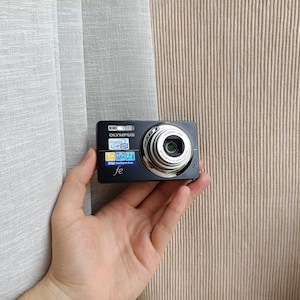 May include: A black Olympus FE digital camera featuring a silver lens barrel, a built-in flash, and a front-facing display area with colorful product specification stickers including 5x zoom, 2.7 inch screen, and Intelligent Auto branding.