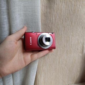 以下が含まれることがあります： 銀色のレンズとフラッシュを備えた赤いCanon PowerShotデジタルカメラ。カメラは手で持たれています。カメラにはCanonのロゴとPowerShotのモデル名が印刷されています。背景はニュートラルです。