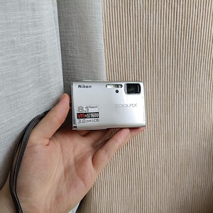 Puede incluir: Una cámara digital Nikon Coolpix plateada con sensor de 8,1 megapíxeles, estabilización de imagen VR, capacidad ISO 1600 y pantalla LCD de 3,0 pulgadas. El dispositivo compacto incluye una correa de muñeca negra unida al cuerpo metálico.