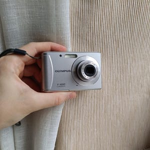 May include: Silver Olympus T-100 digital camera with a black lens and a wrist strap. The camera has a flash and the Olympus logo is visible. The camera is held in a hand.