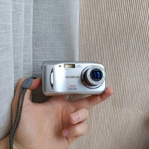 May include: A silver Olympus C-480 ZOOM digital camera featuring a 4.0 megapixel lens, a built-in flash, and a grey wrist strap. The front of the device displays the brand name, the model number, and technical specifications printed in black and red.