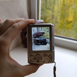 May include: A silver Nikon digital camera with a rectangular body, a rear LCD screen displaying a photograph of a vintage film camera in a brown leather case, a multi-directional navigation pad, and a wrist strap attached to the side.